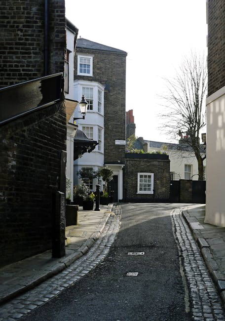 A narrow residential street in Fortune Green and Iverson Road, featuring a cobblestone-edged asphalt road curving gently to the right. The scene includes multi-story brick houses with white-framed sas